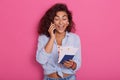 Studio shpt of attractive young woman with dark curly hair, speaking on smartphone with excited facial expression, telling about