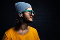 Studio side portrait of young confident man with wireless earphones wearing yellow t-shirt, blue sunglasses and grey hat on black. Royalty Free Stock Photo