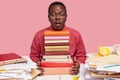 Studying and emotions concept. Puzzled black man poses at desktop near pile of books, papers has stupefied expression