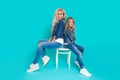 Stunning beauty of a young mother with a cute blonde daughter sitting on a chair on a blue background dressed Royalty Free Stock Photo