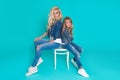 Stunning beauty of a young mother with a cute blonde daughter sitting on a chair on a blue background dressed Royalty Free Stock Photo