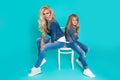 Stunning beauty of a young mother with a cute blonde daughter sitting on a chair on a blue background dressed Royalty Free Stock Photo