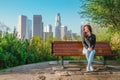 Stunning Brunette Woman In A Denim Jacket Sits On A Park Bench With A View Of Downtown Skyscrapers In Los Angeles