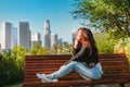 Stunning Brunette Woman In A Denim Jacket Sits On A Park Bench With A View Of Downtown Skyscrapers In Los Angeles