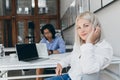 Stunning female programmer playing with blonde hair while posing at workplace with asian male colleague. Busy chinese Royalty Free Stock Photo