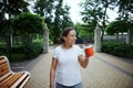 Stunning Latin American young woman holding takeaway coffee in cardboard mug, walking along the alley of a city park