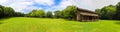 A stunning panoramic shot of a log cabin on a long stretch of lush green grass surrounded by lush green trees with powerful clouds