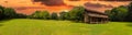 A stunning panoramic shot of a log cabin on a long stretch of lush green grass surrounded by lush green trees with powerful clouds Royalty Free Stock Photo