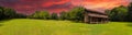 A stunning panoramic shot of a log cabin on a long stretch of lush green grass surrounded by lush green trees with powerful clouds Royalty Free Stock Photo