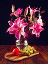 Stunning pink lily bouquet in glass vase on a wooden table and fresh fruit. Still life. Dark background Royalty Free Stock Photo