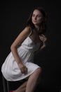 Stunning portrait of a sexy young brunette girl in a white dress sitting on a high chair on a black background. Low key. Beauty Royalty Free Stock Photo