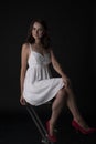 Stunning portrait of a sexy young brunette girl in a white dress sitting on a high chair on a black background. Low key. Beauty Royalty Free Stock Photo