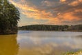 A stunning shot of the vast still waters of a lake surrounded by lush green trees with powerful clouds at sunset