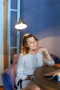 Stunning young girl looks away with downcast eyes, holds hand near face and sits in chair at table on background of lamp