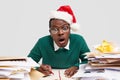 Stupefied dark skinned male opens mouth with amazement, wears spectacles and red festive hat, surprised with many tasks