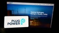 Person holding cellphone with logo of American hydrogen fuel cell company Plug Power Inc. on screen in front of web page.