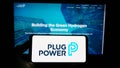 Person holding mobile phone with logo of American hydrogen fuel cell company Plug Power Inc. on screen in front of webpage.
