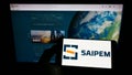 Person holding smartphone with logo of Italian oilfield services company Saipem S.p.A. on screen in front of website.