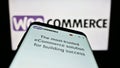 Mobile phone with webpage of open-source e-commerce solution WooCommerce on screen in front of business logo.