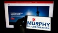 Person holding cellphone with logo of American exploration company Murphy Oil Corporation on screen in front of webpage.