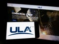 Person holding cellphone with logo of American space company United Launch Alliance (ULA) on screen in front of webpage.