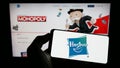 Person holding cellphone with logo of American toys and entertainment company Hasbro Inc. on screen in front of webpage.