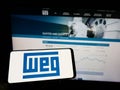Person holding cellphone with logo of Brazilian company WEG Equipamentos Eletricos SA on screen in front of webpage.