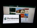 Person holding cellphone with logo of British housebuilding company Persimmon plc on screen in front of business webpage.