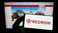Person holding cellphone with logo of British housebuilding company Redrow plc on screen in front of business webpage.