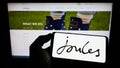 Person holding cellphone with logo of British retail company Joules Group plc on screen in front of business webpage.