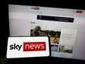 Person holding cellphone with logo of British television channel Sky News on screen in front of business webpage.