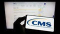 Person holding cellphone with logo of Centers for Medicare and Medicaid Services (CMS) on screen in front of webpage. Royalty Free Stock Photo