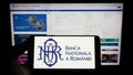 Person holding cellphone with logo of central bank Banca Nationala a Romaniei (BNR) on screen in front of webpage. Royalty Free Stock Photo