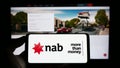 Person holding cellphone with logo of company National Australia Bank Limited (NAB) on screen in front of webpage.