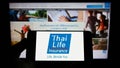 Person holding cellphone with logo of company Thai Life Insurance Public Co. Ltd. on screen in front of business webpage.