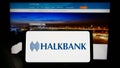 Person holding cellphone with logo of company Turkiye Halk Bankasi A.S. (Halkbank) on screen in front of webpage.