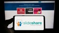 Person holding cellphone with logo of content hosting company SlideShare on screen in front of business webpage.