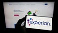 Person holding cellphone with logo of credit reporting company Experian plc on screen in front of business webpage.