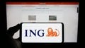 Person holding cellphone with logo of Dutch financial services company ING Groep N.V. on screen in front of webpage.