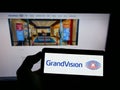 Person holding cellphone with logo of Dutch optical retailing company GrandVision NV on screen in front of webpage.