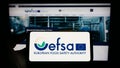 Person holding cellphone with logo of EU agency European Food Safety Authority (EFSA) on screen in front of webpage.