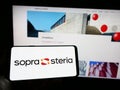 Person holding cellphone with logo of French company Sopra Steria Group SA on screen in front of business webpage.