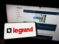 Person holding cellphone with logo of French electronics company Legrand S.A. on screen in front of business webpage.