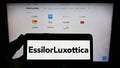 Person holding cellphone with logo of French eyewear company EssilorLuxottica S.A. on screen in front of business webpage. Royalty Free Stock Photo