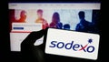 Person holding cellphone with logo of French facility management company Sodexo S.A. on screen in front of webpage.