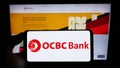 Person holding cellphone with logo of Oversea-Chinese Banking Corporation (OCBC Bank) on screen in front of webpage. Royalty Free Stock Photo