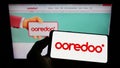 Person holding cellphone with logo of Qatari telecommunications company Ooredoo QSC on screen in front of webpage.