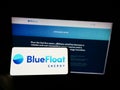 Person holding cellphone with logo of Spanish renewables company BlueFloat Energy LLC on screen in front of webpage.