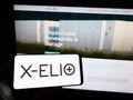 Person holding cellphone with logo of Spanish renewables company X-ELIO Energy SL on screen in front of business webpage.