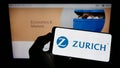 Person holding cellphone with logo of Swiss financial company Zurich Insurance Group AG on screen in front of webpage.